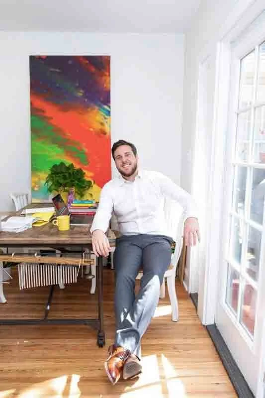 A man sitting on a chair in a bright room, smiling, with a colorful abstract painting, a potted plant, a yellow mug, and books on a wooden desk nearby. There's a large window or glass door to his right letting in sunlight.