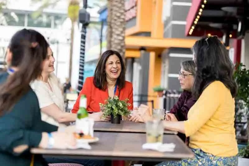 A group of four women sitting at an outdoor table, laughing and talking, with drinks and a small plant in a pot on the table.