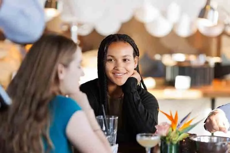 A woman with braided hair smiling and resting her chin on her hand, talking to a woman with long hair in a blue shirt, in a stylish restaurant or bar with warm lighting and decorative elements.