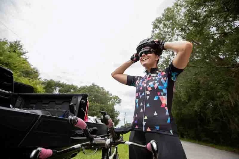 A woman in cycling gear and sunglasses puts on her helmet next to her bicycle on a rural road surrounded by trees.