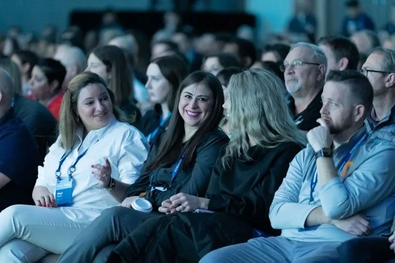 Group of diverse people sitting closely at an indoor conference or seminar, listening attentively and smiling, with some wearing conference badges.