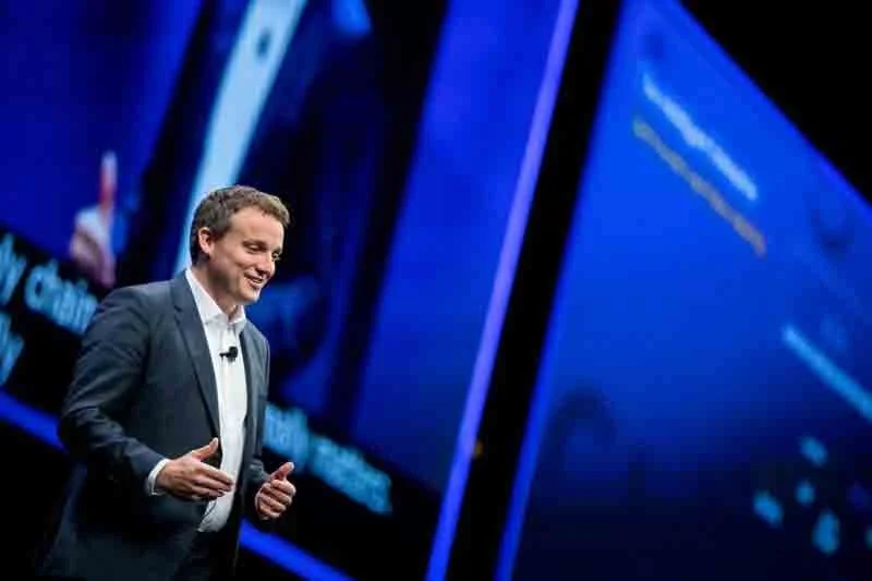 A man in a dark suit giving a presentation on stage with large screens displaying digital content behind him.
