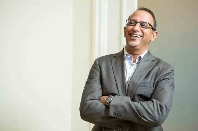 Smiling man in glasses and business suit standing with arms crossed near a window.
