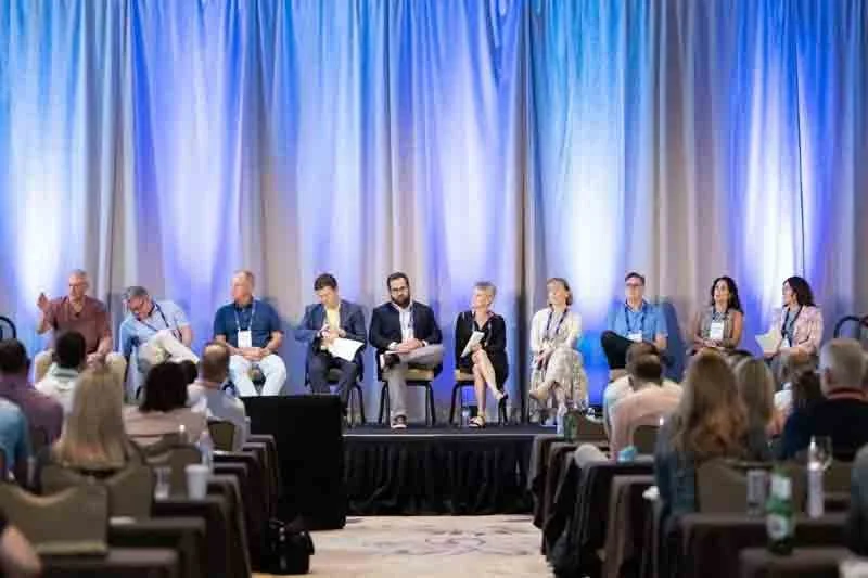 Panel of ten people sitting on a stage in front of an audience at a conference or seminar.