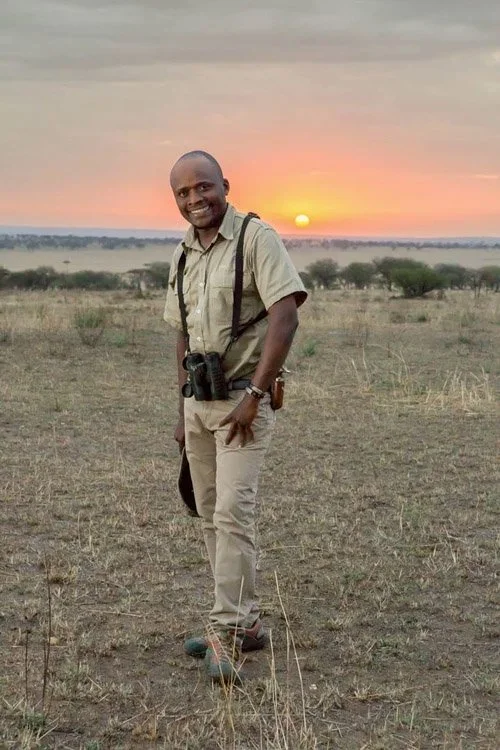 A man outdoors during sunset, dressed in beige safari-style clothing with binoculars hanging around his neck and a camera in his hand, smiling at the camera.