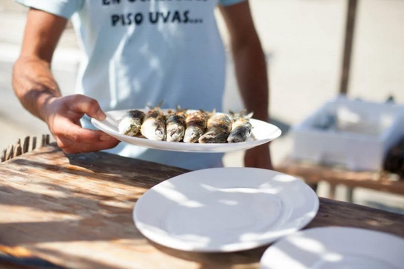 Person holding a white plate with grilled fish, standing outdoors near a table with empty plates.
