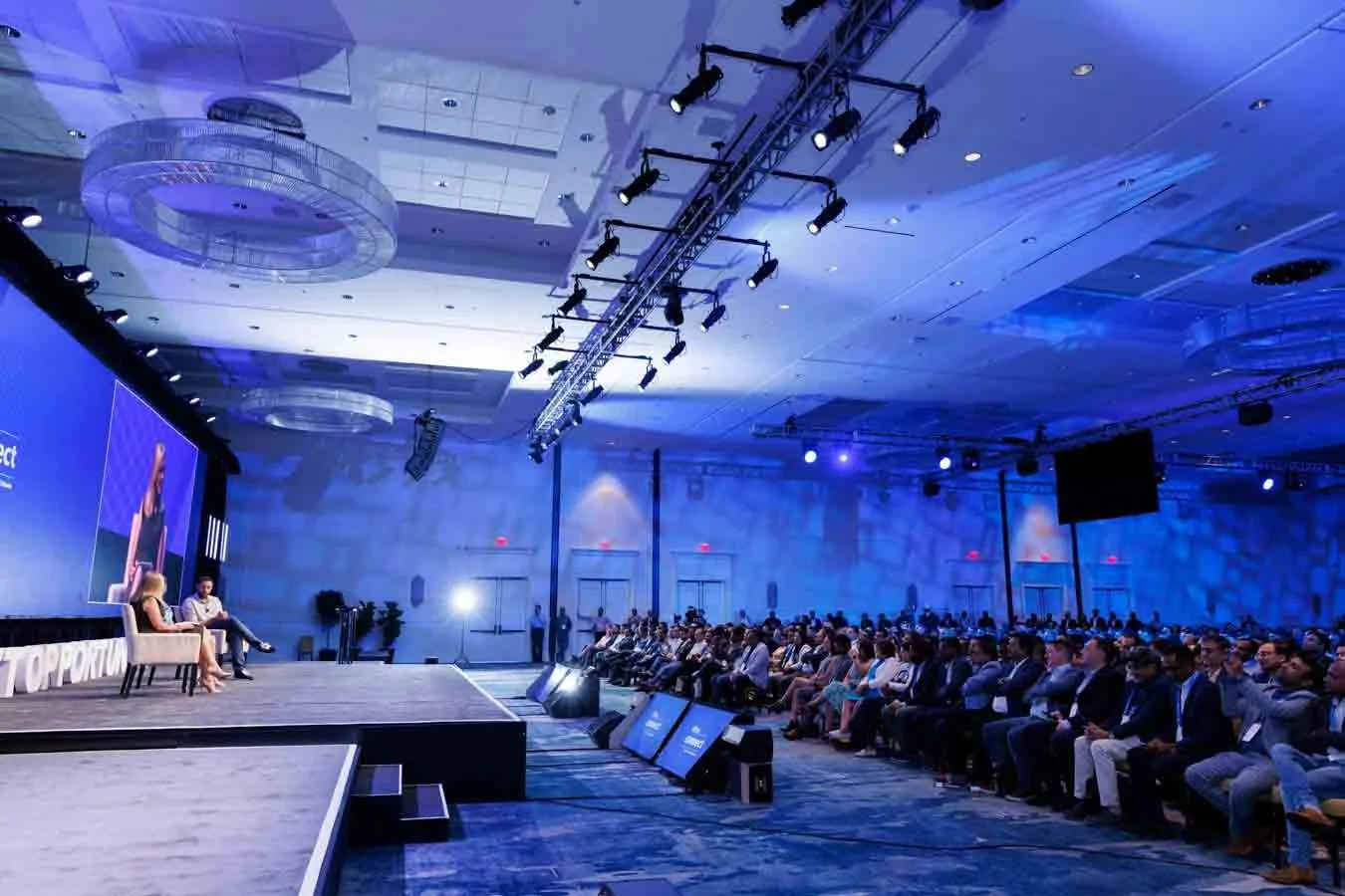 A conference room filled with an audience watching a panel discussion on stage. The stage has three people seated, with a large screen displaying a presentation.