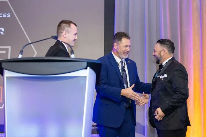 Two men shaking hands on stage at an event, with a third man smiling nearby and a podium with microphones in the foreground.