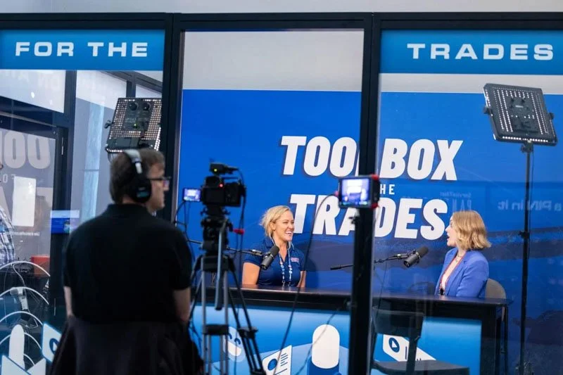 Two women are inside a radio studio or recording booth with blue background and signs reading 'TOO BOX THE TRADES'. They are talking into microphones as part of a broadcast or podcast, with radio equipment and cameras capturing the scene, and a perso
