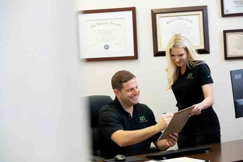 A man sitting at a desk and a woman standing next to him, both smiling, in an office with framed diplomas on the wall.