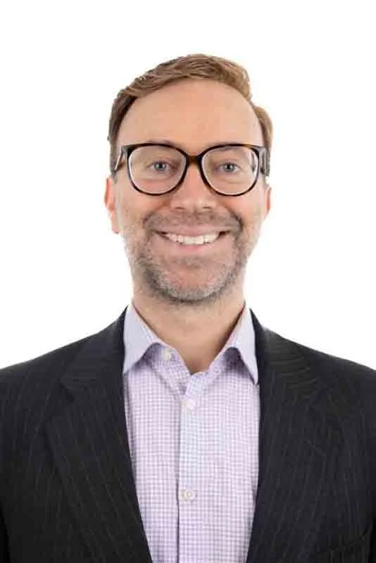 Portrait of a smiling man with glasses, wearing a dark suit and light purple shirt