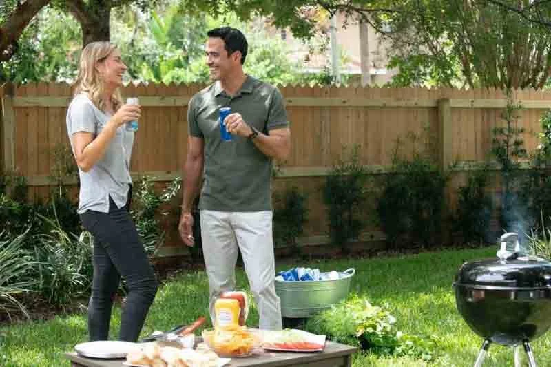 A man and woman talking and smiling in a backyard with a barbecue grill, a table with food, and a wooden fence, surrounded by greenery.