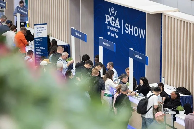 People standing in line at the PGA of America show registration area with signages indicating 'Exhibitors' and 'Purchase Education Passes' in a conference hall.