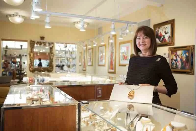 A woman standing inside an art gallery with artwork framed on the wall behind her, and a glass display case filled with jewelry in front of her.