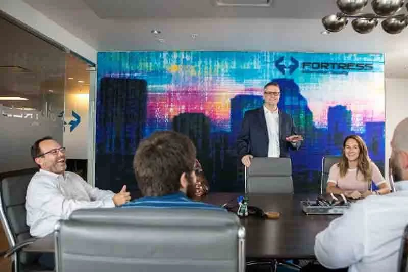Business meeting in a conference room with five people, a man standing and speaking, with a colorful cityscape and 'Fortress' logo in the background.