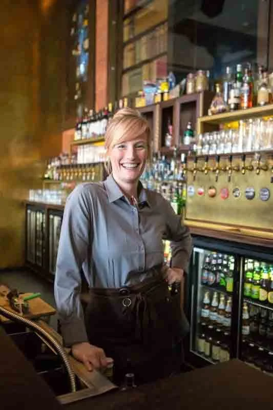 A smiling waitress in a gray shirt standing behind a bar, with beer taps and shelves of bottles in the background.