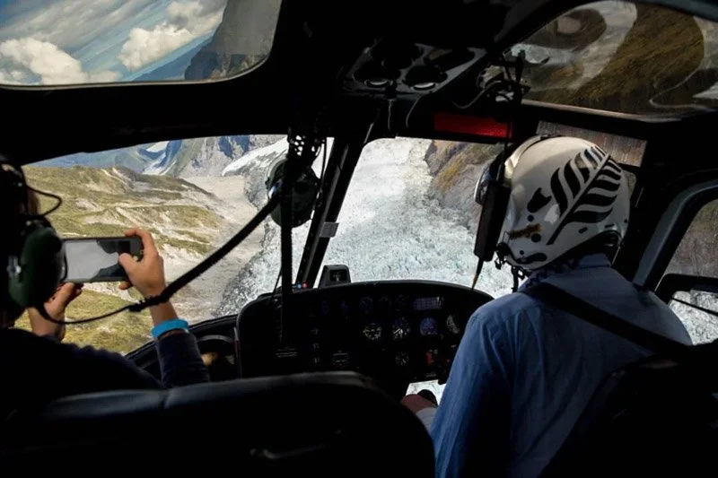 View from inside a helicopter cockpit flying over rocky, mountainous landscape with a glacier, with two people in the front seats, one taking a photo.