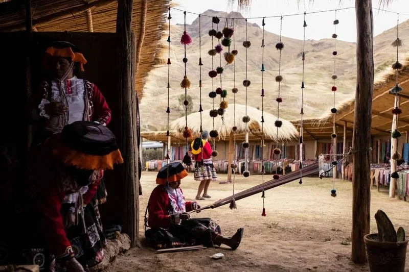 Indigenous women and children in traditional clothing and hats at a cultural site with hanging decorations, wooden structures, and mountainous landscape in the background.