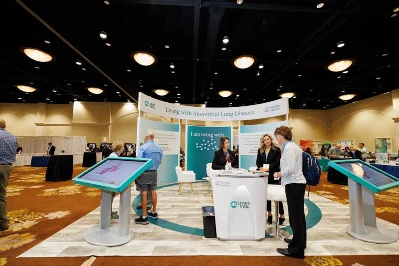 Exhibit booth at a conference with people engaging, discussing about living with interstitial lung disease, featuring informational displays and a seating area.