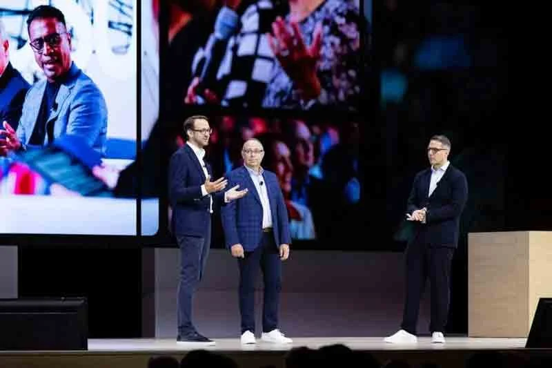Three men standing on a stage in front of a large screen displaying images of people, engaged in a discussion or presentation.