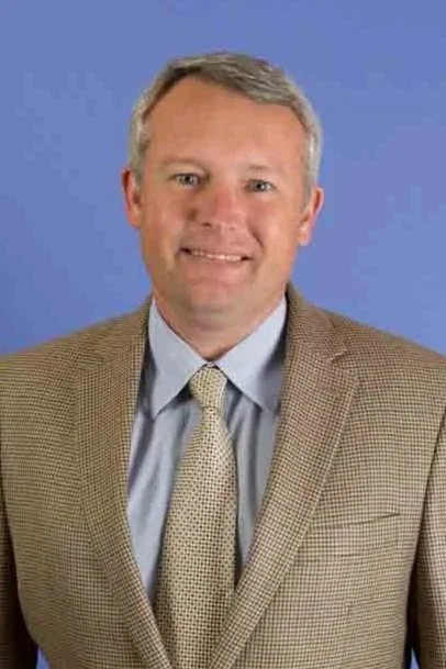 A middle-aged man with gray hair wearing a tan checkered blazer, light blue shirt, and a yellow patterned tie, smiling against a blue background.