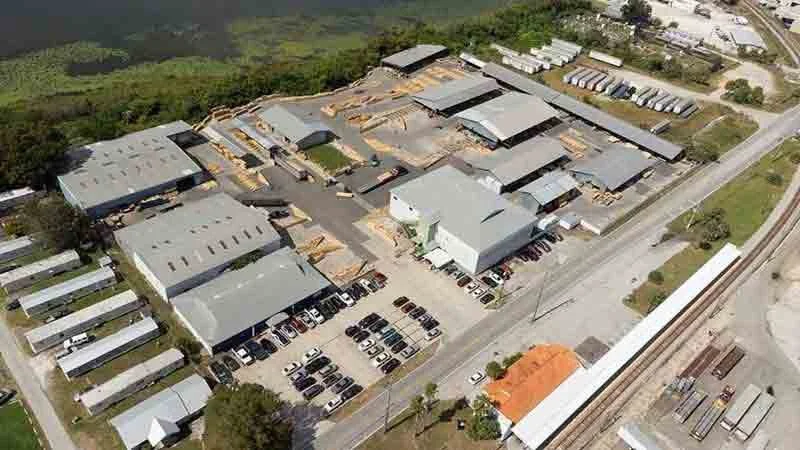 Aerial view of an industrial or warehouse complex with multiple buildings, parking lots filled with cars, and a nearby train track.