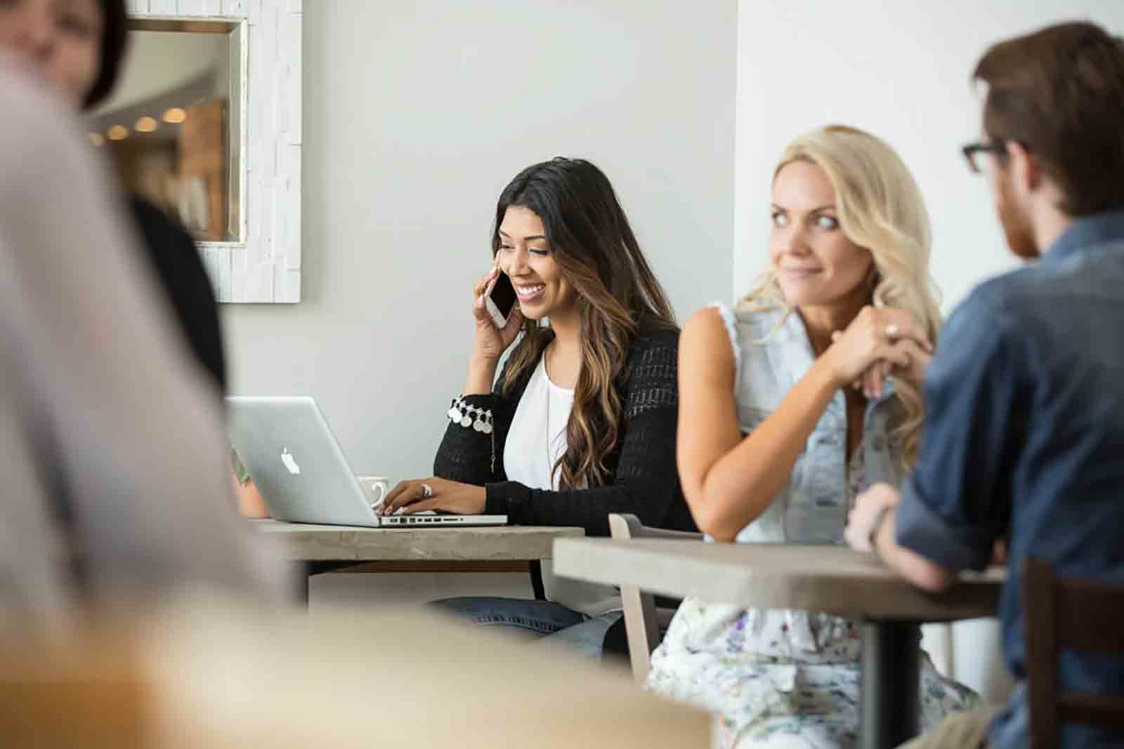 Four people sitting at a table in a casual meeting or co-working space. One woman with long dark hair is talking on her phone and smiling, with a laptop in front of her. Another woman with blonde hair is sitting next to her, looking at a man with glasses across from her. The focus is on the group engaging in conversation.