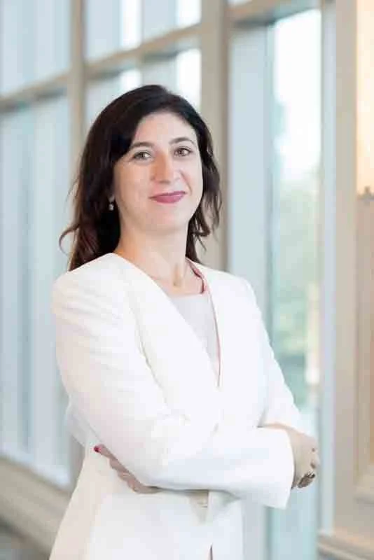 A woman with dark hair, wearing a white blazer, standing with her arms crossed in front of large windows with a view outside.