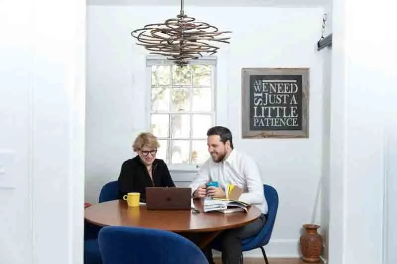 Two people sitting at a round wooden table talking and laughing, with a laptop, coffee mug, and open book, in a bright room with a window and framed sign on the wall that says 'All we need is just a little patience'.