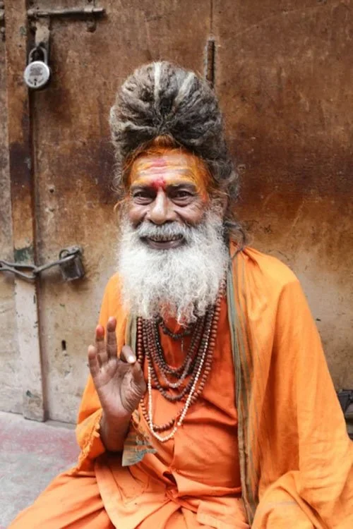 A cheerful man dressed in orange with a long white beard, wearing multiple necklaces, sitting against a rustic wall, raising his hand in a welcoming gesture.