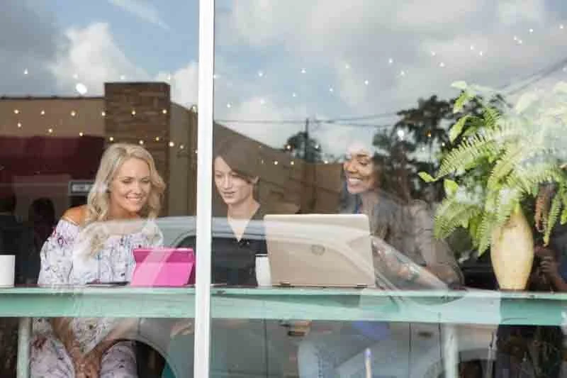 Three women sitting inside a coffee shop, looking out the window and smiling, with a laptop, a tablet, mugs, and plants on the table.