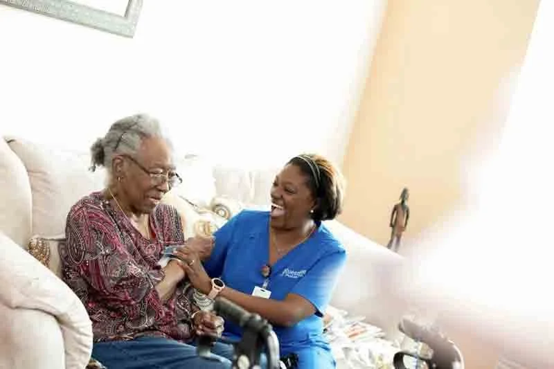 A caregiver and elderly woman sharing a joyful moment in a living room, sitting on a sofa with sunlight streaming in.