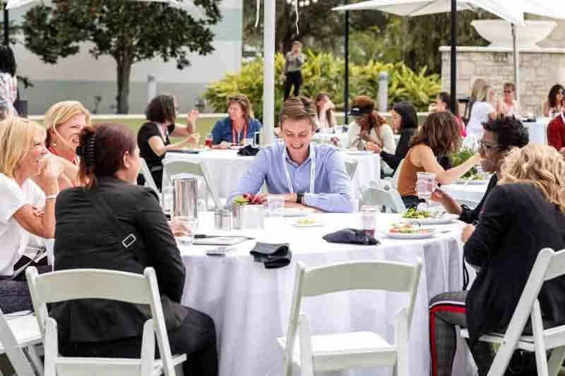 People enjoying a meal outdoors at a garden event with white tables and chairs, and large umbrellas.