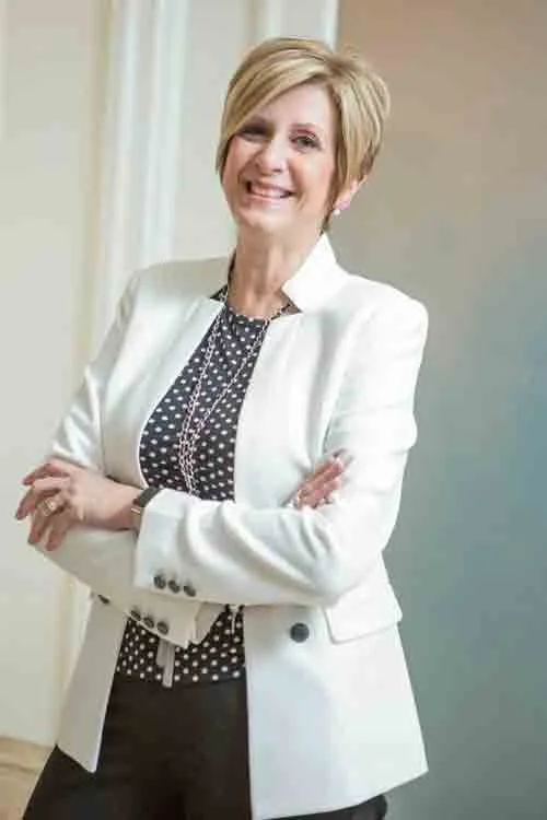 A woman with short blonde hair smiling and standing with arms crossed, wearing a white blazer over a black and white polka dot blouse.