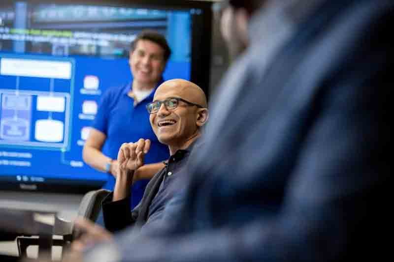 Two men in a meeting room, one wearing a blue shirt and the other wearing glasses and a dark blazer, smiling and engaged in a discussion. A large screen with a blue digital interface is in the background.