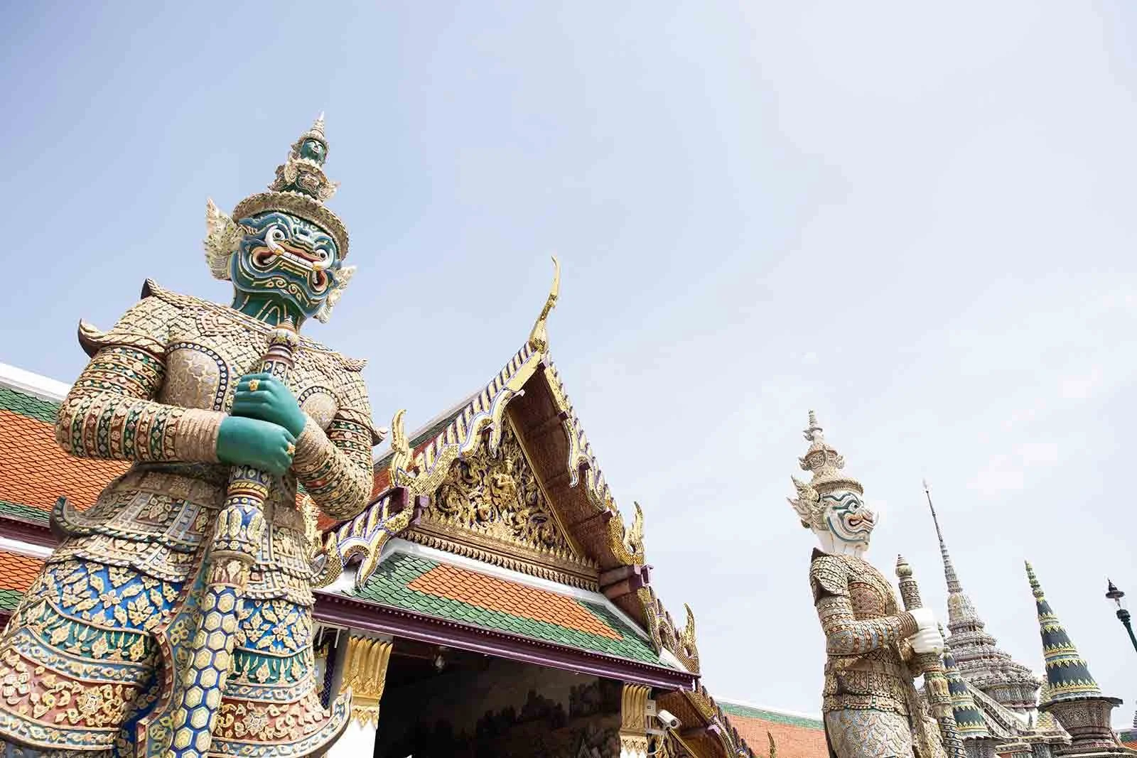 Two large traditional Thai statues with intricate designs and fierce expressions, standing in front of a richly decorated temple with a colorful roof and golden details, against a clear sky.