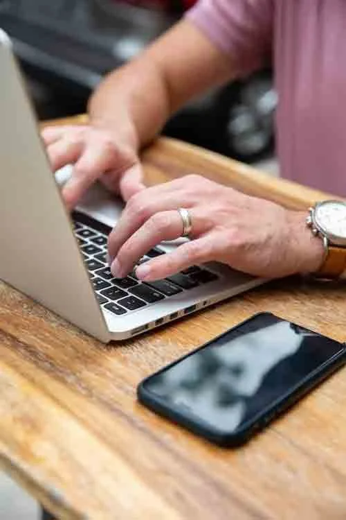 Person typing on a silver laptop with a smartphone on a wooden table.