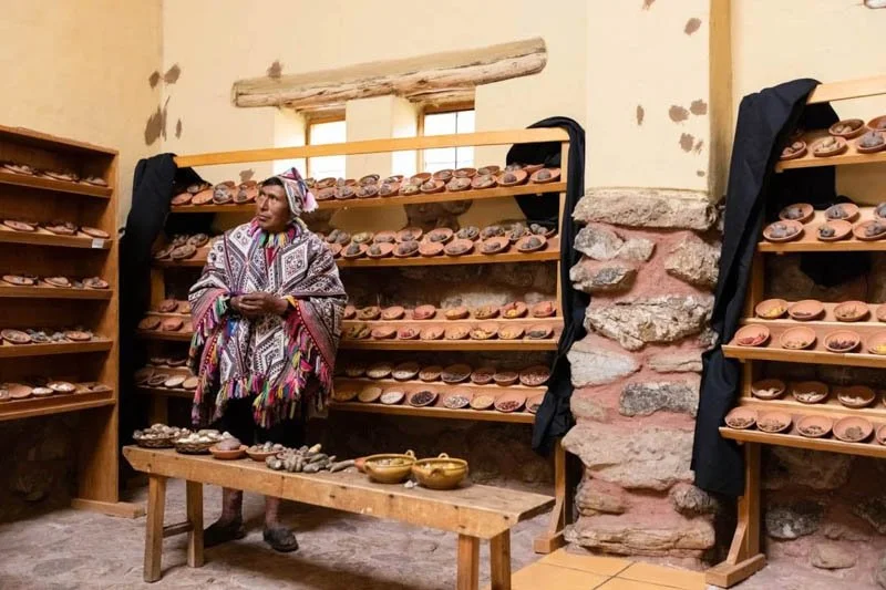 A woman in traditional attire stands in front of wooden shelves filled with clay pots, in a rustic shop or workshop setting with stone and wood walls.