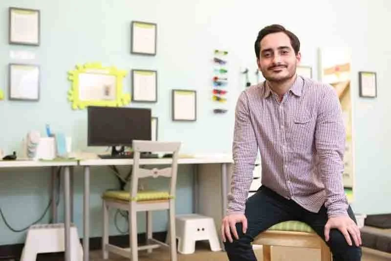 A man sitting on a small stool in an office with a computer desk, monitor, and framed pictures on the wall.