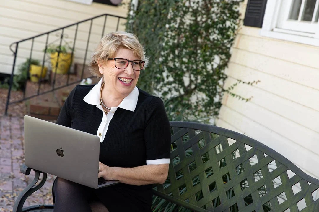 A woman with short blonde hair, glasses, and a black and white blouse sitting on a green metal bench outside, smiling while looking to her left, with a silver Apple laptop on her lap, in front of a beige house with white trim and greenery.