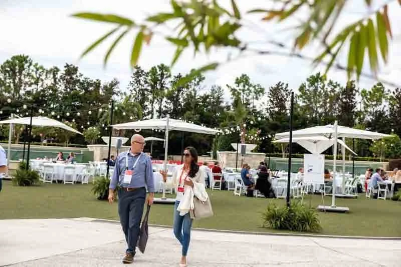 Outdoor event with people walking on pavement, dining under white umbrellas, string lights overhead, grassy area, trees, and clear sky
