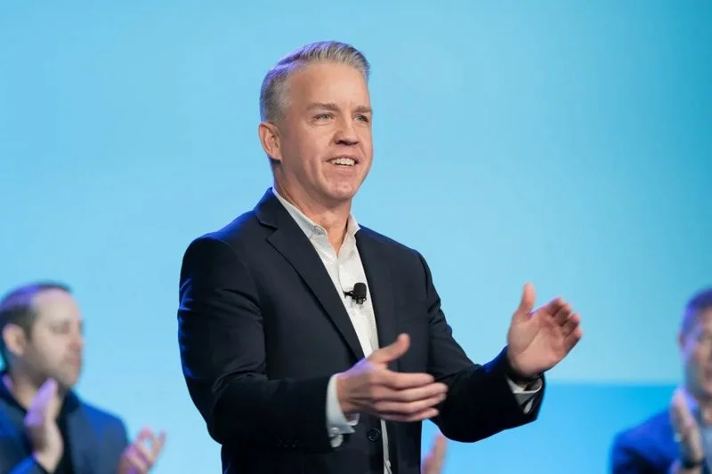 A man in a suit speaking on stage with a light blue background, with several people clapping in the background.