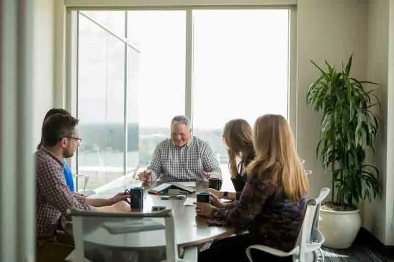 A group of five people sitting around a conference table in a bright room, engaging in a discussion, with laptops and coffee mugs on the table, and large windows in the background.