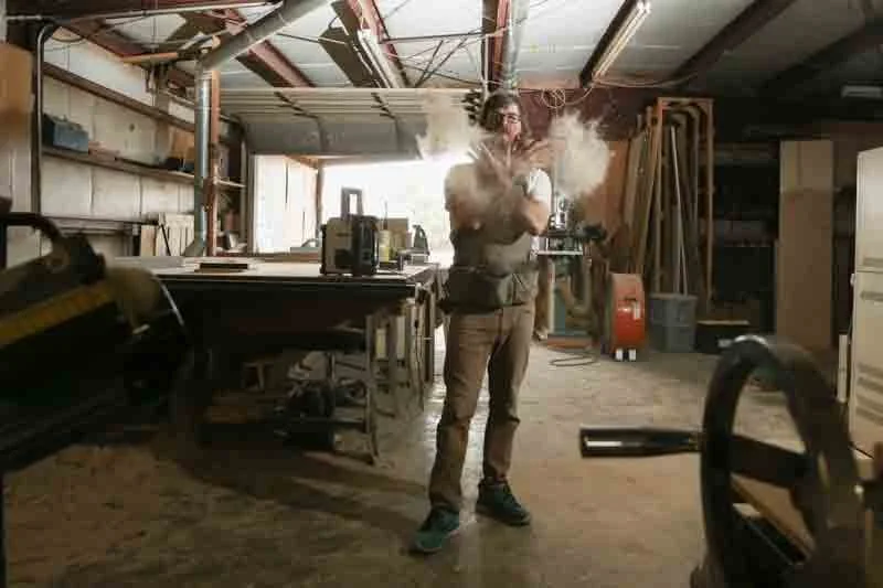 Person standing in a workshop with arms crossed, blowing out smoke or dust, surrounded by woodworking tools and equipment.