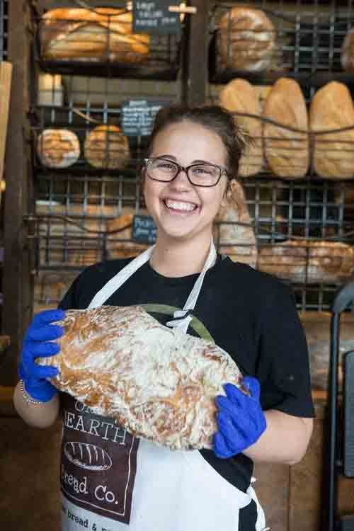 Smiling woman wearing glasses, black shirt, and blue gloves holding a large fish in a bakery with bread on shelves in the background.