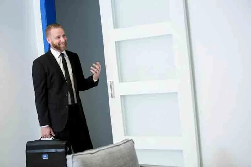 Man in a black suit and tie holding a briefcase, smiling, standing near a door in a modern office space.