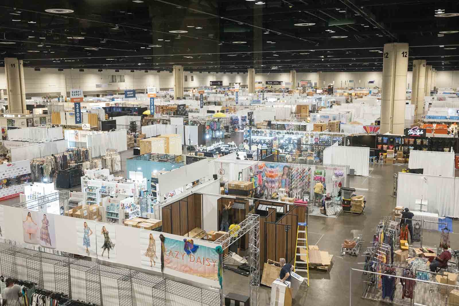 Large indoor convention center with multiple booths selling clothing, accessories, and other items. White partition walls and signs indicating booth numbers. Several people browsing and shopping.