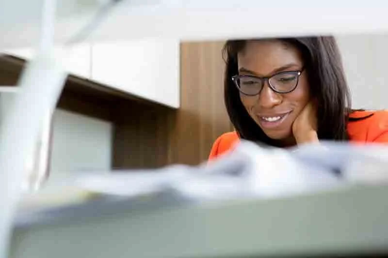 A woman with glasses smiling while looking at papers on her desk in an office setting.