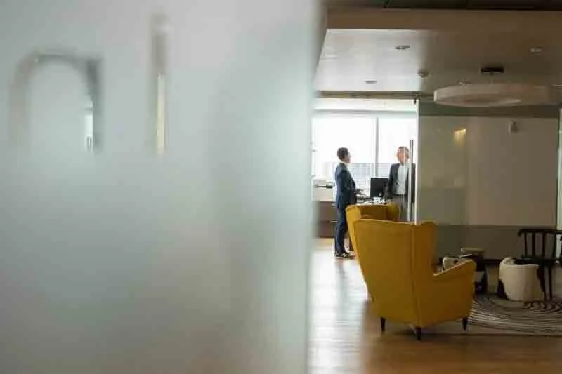 Two people in business attire having a conversation in a modern office lounge area with yellow armchairs and a large window in the background.
