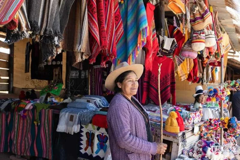 Woman with a hat standing in front of a market stall selling colorful textiles and handicrafts in a traditional market setting.